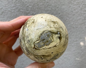 May include: A spherical, decorative stone ball with a marbled pattern in shades of beige, gray, and olive green. The stone has a rough, textured surface and a unique, organic appearance. The ball is held in a hand.