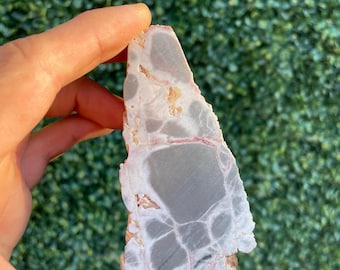 May include: A polished, triangular-shaped mineral specimen featuring a smooth, flat surface with a mottled pattern of soft pink, light gray, and white tones. The stone displays distinct veining and irregular patches of color throughout its structure.