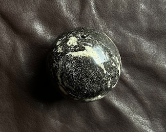 May include: A polished, spherical stone with a speckled black and white pattern. The orb is smooth and reflective, with a glossy finish. The stone is set against a dark brown, textured background.