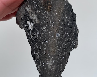 May include: A dark gray, irregularly shaped rock specimen featuring a rough, textured surface with scattered white and light gray mineral inclusions throughout. The stone has a pointed, triangular form with visible crystalline flecks and dark patches.