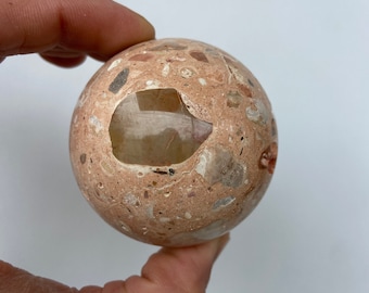 May include: A spherical, speckled stone object with a large, irregular opening. The sphere is primarily a light peach color, with embedded fragments of various colors, including gray, white, and red. The object is held in a hand against a white background.