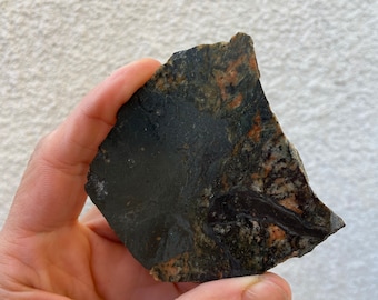 May include: A dark, irregularly shaped rock with a smooth, black surface and areas of orange and speckled patterns. The rock is held in a hand, showcasing its natural textures and colors.