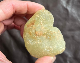 May include: A light green, irregularly shaped stone with a rough texture and brown markings. The stone is held in a hand, showcasing its natural form and color variations. The stone appears to be a raw, uncut gemstone.