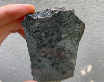 May include: A dark gray, irregularly shaped stone with a rough, textured surface. The stone has a mix of dark and light gray areas, with some areas appearing almost black. The edges are jagged and uneven, giving it a natural, raw appearance.