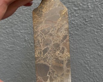 May include: A rectangular, polished stone with a marbled pattern of brown, tan, and gray. The stone is held by a hand, showcasing its natural texture and unique veining. The top edge is angled, and the bottom edge is uneven.