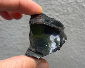 May include: A piece of black obsidian, a volcanic glass, held between fingers. The stone has a glossy, reflective surface with visible fractures and a dark, opaque color. The edges are rough and uneven.