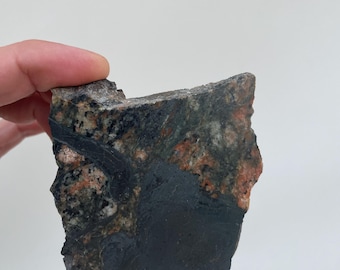 May include: A flat, irregularly shaped slab of natural stone featuring a mottled texture with prominent patches of dark charcoal gray, speckled black, white, and earthy orange-red mineral inclusions throughout the rough, unpolished surface.