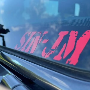 May include: A red vinyl sticker with the words "SIN-EM" in a bold, stylized font. The sticker is applied to the rear window of a vehicle.