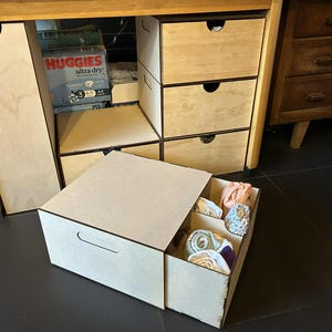 May include: A light-colored wooden storage box with a lid and handle, divided into compartments. The box contains folded fabric items. Other wooden storage units and a desk are in the background.