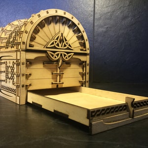 May include: A wooden box with a Celtic knot design on the lid and a drawer that slides out. The box is made of light brown wood and has a natural finish.