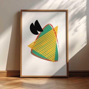 May include: A framed abstract art print with a geometric design. The artwork features a green triangle with yellow stripes, a yellow triangle, red accents, and black shapes. The print is in a wooden frame and leans against a white wall.