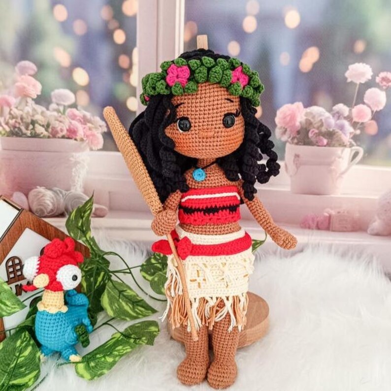 Op de afbeelding: Een gehaakte pop met een bruine huid, zwart krullend haar en een bloemenkroon. Ze draagt een rood-wit topje, een beige rok met franjes en een bruine stok. De pop staat op een houten voet.