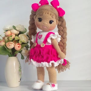 May include: A crocheted doll with long brown braids, wearing a pink and white dress with a heart-shaped design. The doll has a pink bow in her hair and is holding a pink flower.