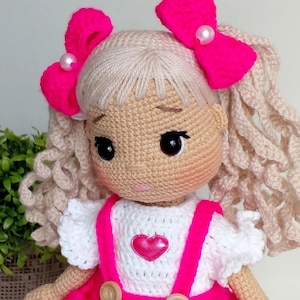 May include: A crocheted doll with blonde curly hair, wearing a white shirt with pink straps and a pink skirt. The doll has pink bows in her hair and a pink heart on her shirt.