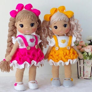 May include: Two crocheted dolls with long hair, one wearing a pink dress and the other wearing a yellow dress. Both dolls have white crocheted tops and are wearing crocheted shoes.