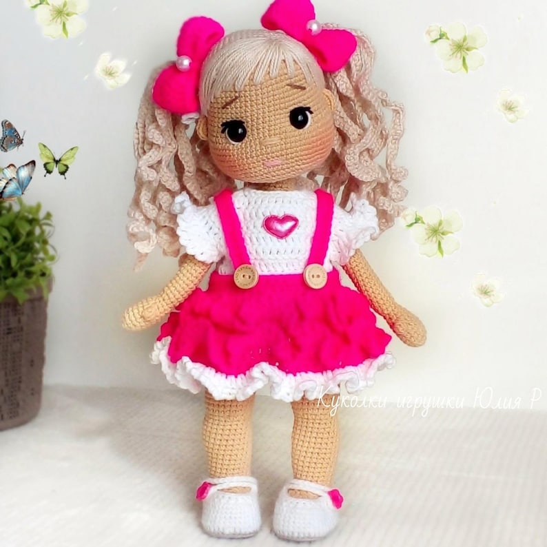 May include: A crocheted doll with blonde curly hair, wearing a pink dress with white trim and a white shirt with a heart on the front. The doll has pink bows in her hair and is wearing white shoes.