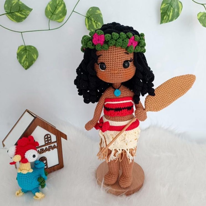 Op de afbeelding: Een gehaakte pop van Moana uit de Disney-film Vaiana. De pop draagt een rood-wit topje, een bruin-wit rokje en een bloemenkrans. Ze houdt een bruin houten peddel vast.