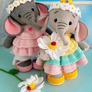 Puede incluir: Dos juguetes de elefante de crochet que llevan vestidos y coronas de flores. El elefante de la izquierda lleva un vestido rosa y el elefante de la derecha lleva un vestido amarillo, verde y rosa. Ambos elefantes sostienen flores blancas de crochet.