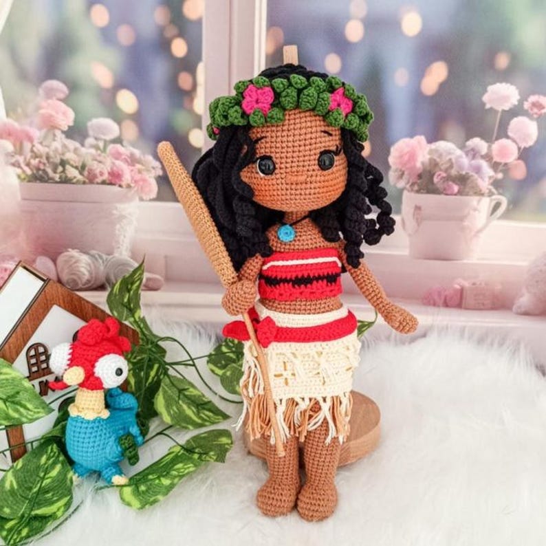 Op de afbeelding: Een gehaakte pop met een bruine huid, zwart haar en een bloemenkroon. Ze draagt een rood-zwart topje, een witte rok en een blauwe ketting. Ze houdt een houten stok in haar rechterhand.