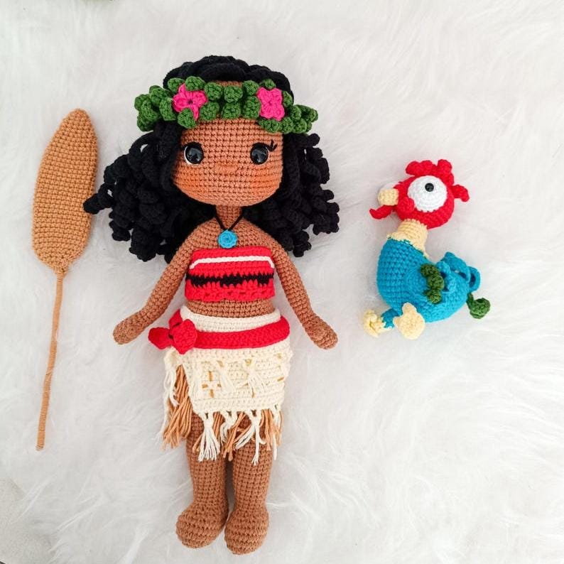 Op de afbeelding: Een gehaakte pop met bruine huid, zwart krullend haar en een bloemenkrans. Ze draagt een rood-wit topje en een beige rok met franjes. Ze houdt een bruine stok vast. Een blauwe en gele gehaakte haan bevindt zich aan de rechterkant van de afbeelding.