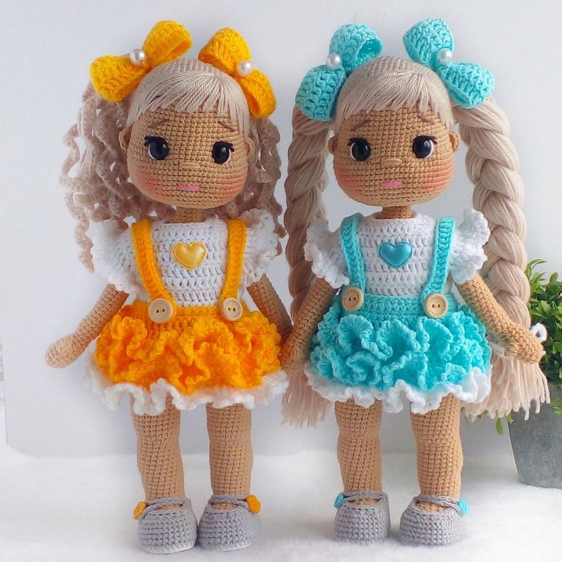 May include: Two crocheted dolls with blonde hair and braided pigtails. One doll is wearing a yellow dress with white straps and a white heart on the front. The other doll is wearing a blue dress with white straps and a white heart on the front.