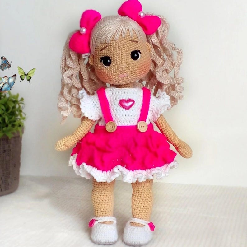 May include: A crocheted doll with blonde curly hair, wearing a white shirt with pink overalls, a pink skirt with white lace trim, and pink bows. The doll is wearing white shoes.