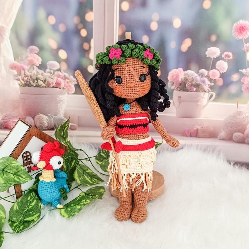 Op de afbeelding: Een gehaakte pop met bruine huid, zwart haar en een bloemenkroon. Ze draagt een rood-wit topje en een witte rok met bruine details. Ze houdt een houten stok vast. Op de achtergrond is een kleine gehaakte vogel te zien.
