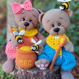 Pode incluir: Dois ursinhos de crochê, um com laço e vestido rosa, o outro com calças azuis e top amarelo. Ambos têm detalhes de abelhas. Há também um pequeno pote laranja com a palavra "Mel" em cirílico e um objeto em forma de pata.