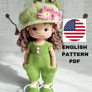 Pode incluir: Padrão de boneca de crochê em inglês. A boneca está vestindo um macacão verde, um chapéu verde com uma flor rosa e cabelo castanho encaracolado. O padrão está disponível para download em PDF.