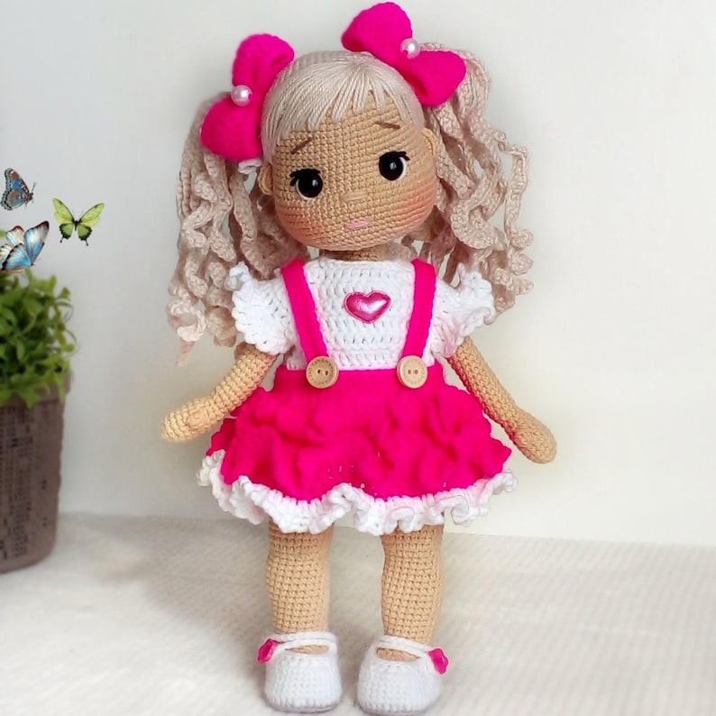 May include: A crocheted doll with blonde curly hair, wearing a pink dress with white trim, white crocheted top with a pink heart, and pink bows in her hair.