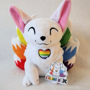May include: White plush fox with a rainbow heart on its chest and a rainbow tail. The fox is sitting on a white surface. The tag says "Pride Fox".