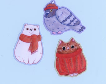 Op de afbeelding: Drie geïllustreerde stickers met een witte ijsbeer met een rode sjaal, een grijze duif met een rode hoed en een bruine kat met een rode trui.