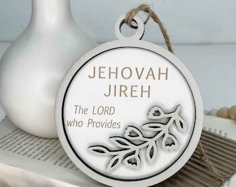 May include: White wooden ornament with a brown twine hanger. The ornament is round and features a floral design with the text "Jehovah Jireh The LORD who Provides".