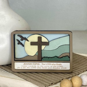 May include: A wooden sign with a brown background and a light blue sky. The sign features a cross, a yellow sun, and two birds flying. The text on the sign reads "JEHOVAH RAPHA - The LORD who Heals Listen carefully to the voice of the LORD, for I am the LORD who heals you... Exodus 15:26"