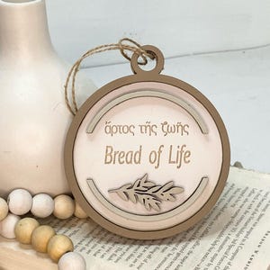 May include: A wooden ornament with a light brown background and a white circle in the center. The circle has a light brown border and the text "ἄρτος τῆς ζωῆς" and "Bread of Life" in brown lettering. A small brown leaf design is in the bottom center of the circle.