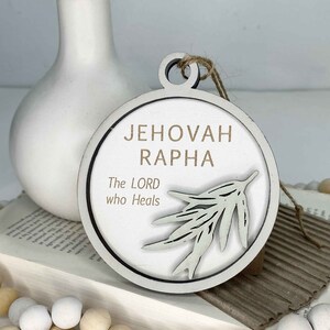 May include: White wooden ornament with a brown circle around the edge. The ornament has a white branch design and the text "Jehovah Rapha The LORD who Heals".
