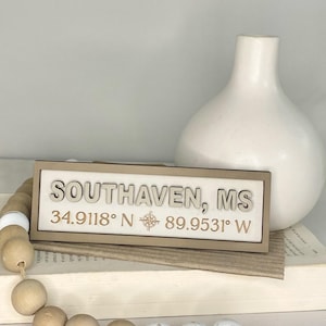 May include: A wooden sign with the text "SOUTHAVEN, MS" and coordinates. The sign is light-colored with a brown frame and a compass rose symbol. A white vase and a wooden bead garland are also in the image.