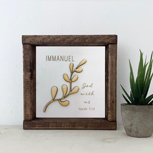 May include: A wooden framed sign with the words "Immanuel God with us Isaiah 7:14" and a gold leaf branch design.