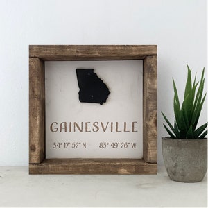 May include: A wooden framed sign with a white background featuring a black silhouette of the state of Georgia and the text "GAINESVILLE 34° 17' 52" N 83° 49' 26" W".