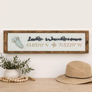 May include: A rectangular wooden sign with a dark brown frame and a light-colored background. It features a cutout map of Lake Waukewan, the text "Lake Waukewan", a compass rose icon, and the coordinates "43.6570° N 71.5229° W" in black and gold.