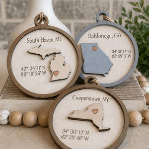 May include: Three wooden ornaments with state outlines and heart details. Each ornament features a different location: South Haven, MI, Dahlonega, GA, and Cooperstown, NY, with coordinates. The ornaments have a circular design with a neutral color scheme.