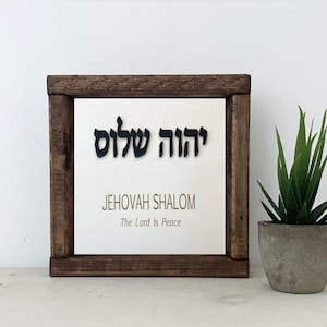 May include: A square wooden sign with a dark brown frame. The sign has a white background with the words "יהוה שלום" in black, above the English text "JEHOVAH SHALOM" and "The Lord Is Peace" in a light brown color.