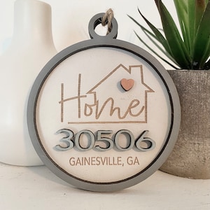 May include: A round wooden ornament with a gray border and a white background. The ornament features a house with a heart in the window and the text "Home" above the house. The number "30506" is below the house, and the text "GAINESVILLE, GA" is below the number.