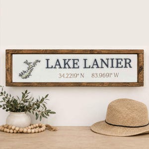 May include: A rectangular wooden sign with a dark brown frame and a white center panel. The sign features a dark blue map outline of Lake Lanier, the text LAKE LANIER in dark blue, and the coordinates 34.2219 N 83.9691 W in dark brown.