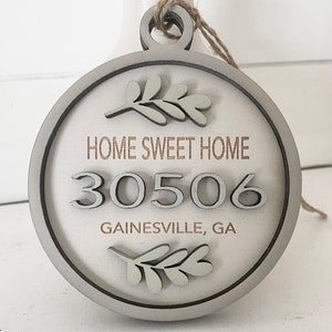 May include: A round, white wooden ornament with a brown rope hanger. The ornament has a gray border and features the text "Home Sweet Home", the number "30506", and the text "Gainesville, GA". The ornament is decorated with two green leaf sprigs.