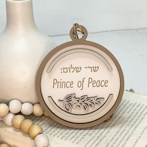 May include: A wooden ornament with a light brown background and a white inner circle. The ornament features the Hebrew words "שר שלום" and the English words "Prince of Peace" in a brown font. A stylized olive branch is depicted in the center of the ornament.