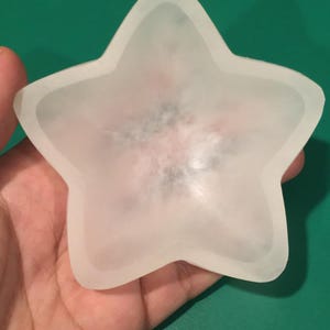 Könnte beinhalten: Eine sternförmige Schale aus mattiertem Glas. Die Schale hat einen leicht erhöhten Rand und ein strukturiertes Inneres. Die Sternschale wird in einer Hand vor einem grünen Hintergrund gehalten. Diese dekorative Schale könnte für Schmuck oder kleine Gegenstände verwendet werden.