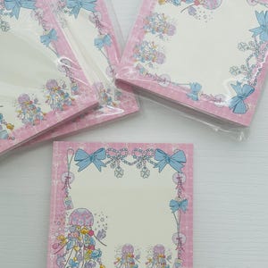 May include: Pink memo pads with a decorative border featuring blue bows, jellyfish, and floral elements. The pads are packaged in clear plastic and have a white interior for writing notes. The design has a whimsical, playful aesthetic.