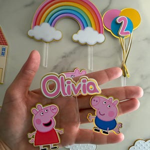 May include: Cake toppers featuring a rainbow, clouds, and balloons with the number 2. Includes Peppa Pig and George Pig characters, and a pink and gold name topper that says "Olivia".