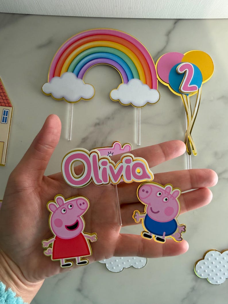 May include: Cake toppers featuring a rainbow, clouds, and balloons with the number 2. Includes Peppa Pig and George Pig characters, and a pink and gold name topper that says "Olivia".